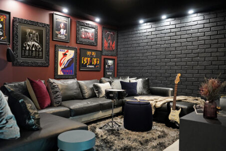 Rock 'n' Roll Inspired Media Room With Curated Art, Deep Maroon Accent Wall, Black Brickwork, And Lounge Sofa, And Musical Instruments.